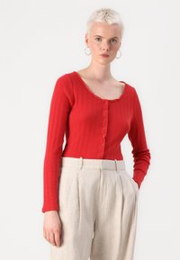 Haut rouge à manches longues côtelées avec un col en cœur, fermeture à boutons et bord festonné. Assorti à un pantalon beige clair taille haute.