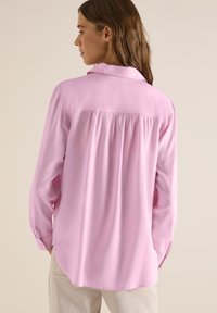 Femme portant un chemisier rose ample à manches longues avec un détail froncé au dos, associé à un pantalon beige clair, vue de profil sur un fond uni.