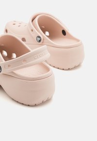 Des Crocs rose clair avec une semelle épaisse, des trous de ventilation et une sangle ajustable. La surface est lisse avec une finition mate et une semelle intérieure texturée.