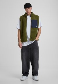 Fleece-Weste in Olivgrün mit einer marineblauen Brusttasche, Frontreißverschluss und Seitentaschen, über einem weißen T-Shirt getragen und mit grauen Jeans kombiniert.