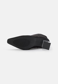 Botte noire en daim à bout pointu avec une semelle texturée et un talon carré à motif, vue du dessous.