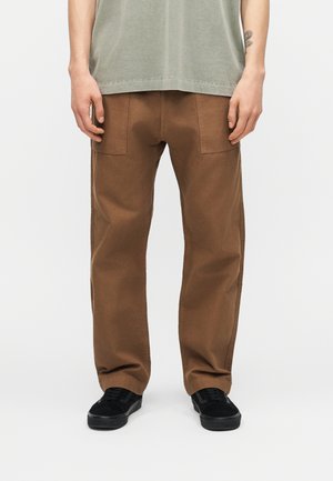 Man draagt bruine broek met rechte pijpen, zwarte sneakers en een grijs shirt, staand tegen een simpele lichte achtergrond.