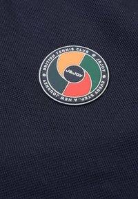 Marineblauwe textiel met een cirkelvormig, meerkleurig embleem waarop "J&JOY" en "BRITISH TENNIS CLUB" is ingeschreven.