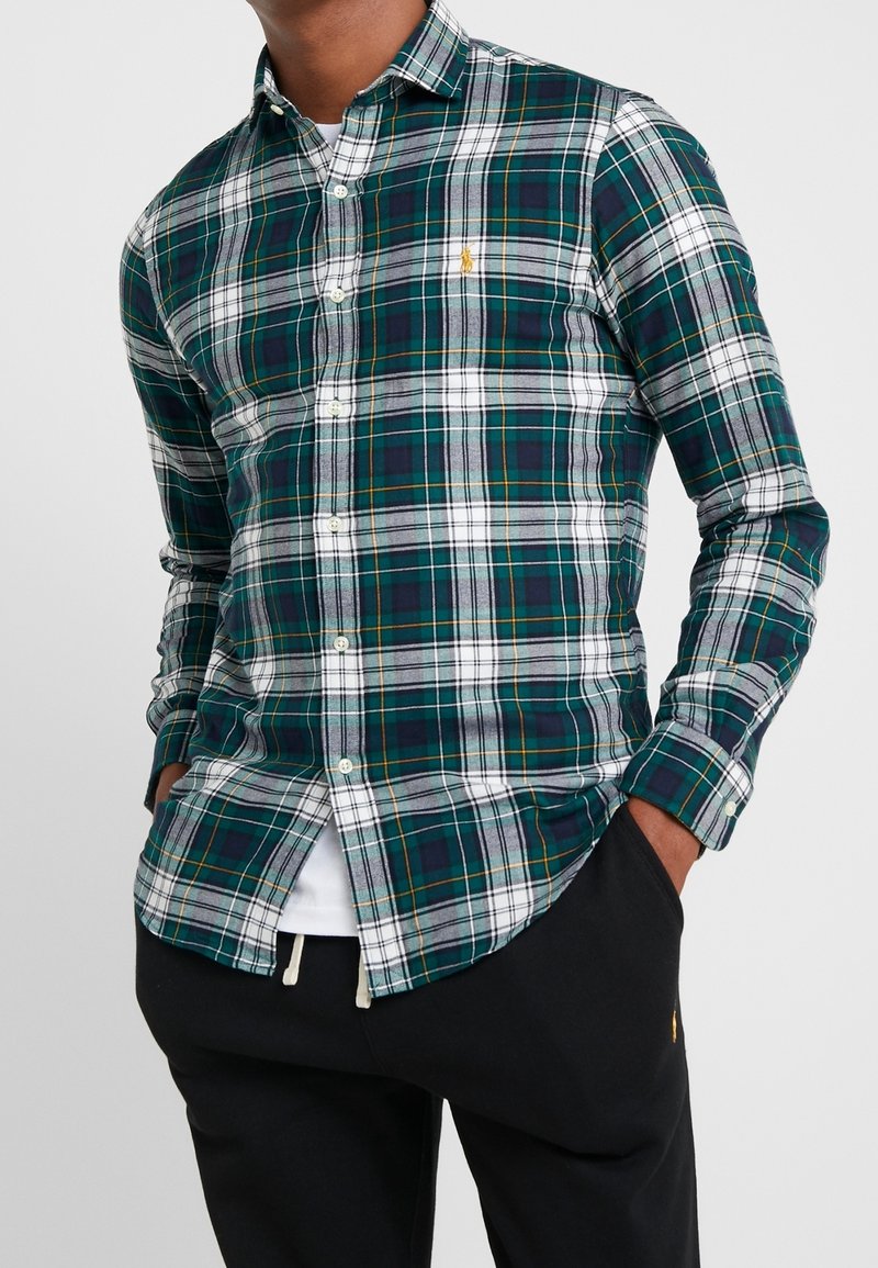 Zielona i biała koszula w kratę z długim rękawem, z kołnierzykiem na guziki oraz akcentem w postaci logo. Materiał wygląda na gładki, a krój jest luźny.