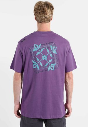 Homme aux cheveux blonds courts portant un t-shirt violet avec un motif floral bleu et du texte au dos, se tenant devant un fond blanc.