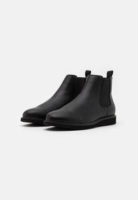 Un par de botas Chelsea negras de piel con paneles laterales elásticos y lengüetas, mostradas sobre un fondo blanco liso.