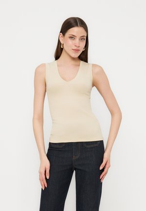 Νέα γυναίκα με μπεζ αμάνικο τοπ με V λαιμόκοψη και σκουρό μπλε τζιν, στέκεται μπροστά σε απλό ανοιχτό φόντο.