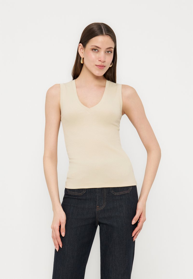 Νέα γυναίκα με μπεζ αμάνικο τοπ με V λαιμόκοψη και σκουρό μπλε τζιν, στέκεται μπροστά σε απλό ανοιχτό φόντο.