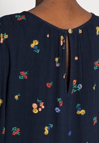 Donkerblauwe blouse met meerkleurige bloemrijke borduursels, een geplooide textuur en een kleine knoopsluiting aan de achterkant van de hals.