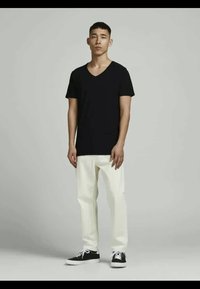 Schwarzes V-Ausschnitt-T-Shirt kombiniert mit weißen Hosen und schwarzen Sneakers. Das Modell steht vor einem hellgrauen Hintergrund. Einfaches und modernes Design.