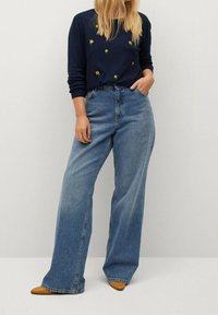 Suéter azul marino oscuro con bordado de flores, combinado con pantalones vaqueros de pierna ancha color azul claro y planos de puntera afilada en tono marrón. Corte relajado, estilo casual.
