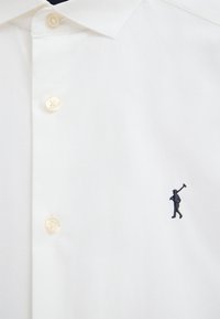 Camisa blanca abotonada con un pequeño logo bordado de una persona llevando un palo de golf en el lado izquierdo cerca de los botones.