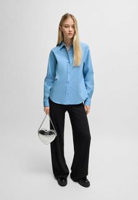 Camicia button-up azzurro chiaro, maniche lunghe, pantaloni neri a gamba larga e una borsa a forma di cuore in argento con tracolla a catena.