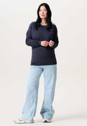 Woman with long black hair wearing a navy sweatshirt, light blue wide-leg jeans, and white sneakers standing with hands clasped.