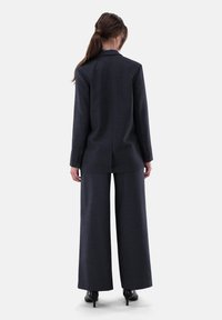 Dunkel maßgeschneiderter Anzug mit weit geschnittenen Hosen und einem taillierten Blazer. Glatte Textur und einfarbig, mit einem dezenten Muster. Schwarze High Heels.