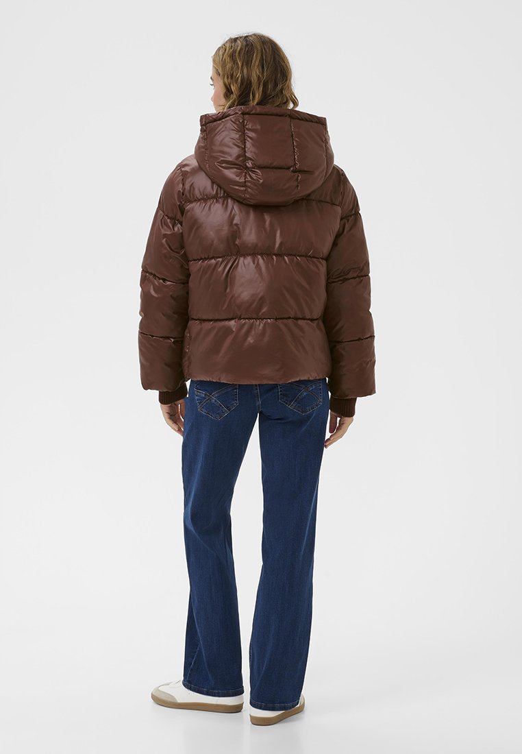 Person, der eine braune, mit Kapuze versehene Steppjacke, blaue Jeans und weiße Sneakers trägt, steht mit dem Rücken zu einer einfarbigen Hintergrund.