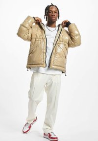 Chaqueta acolchada beige con un acabado brillante, cremallera negra y dos bolsillos frontales, combinada con pantalones blancos y zapatillas rojas y blancas.