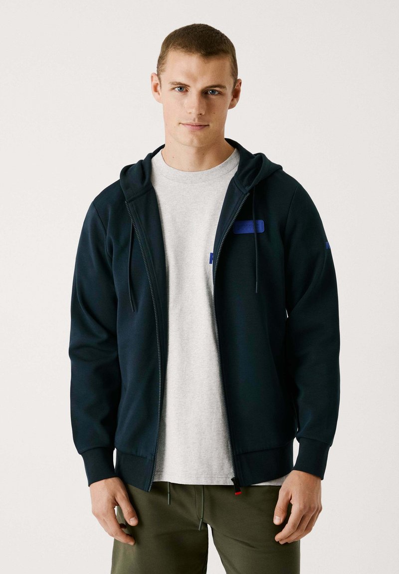 Marinefarbener Reißverschluss-Hoodie mit einer Kordelzugkapuze, mit einem blauen Logo-Patch auf der Brust, getragen über einem hellgrauen T-Shirt, kombiniert mit olivfarbenen Hosen.