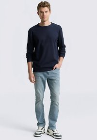 Navy blue long-sleeve sweatshirt with round neck, paired with light blue denim jeans and black/white sneakers. Features subtle stitching details.