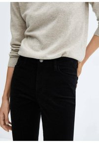 Personne portant un pull beige à manches longues rentré dans un pantalon noir taille haute, une main dans la poche arrière, sur un fond clair uni.