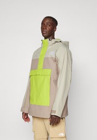 Vindjakke med et design i beige og lys grønn fargeblokk, en frontlomme og justerbar hette med glidelås.