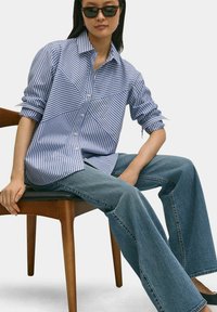 Blauw en wit gestreepte button-up shirt met een geometrisch ontwerp, voorzien van opgerolde mouwen. Gecombineerd met lichtblauwe wijde jeans.