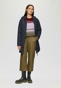 Gevoerde marineblauwe jas met een capuchon, gelaagd over een gestreepte trui in bordeaux, lavendel en groen, gecombineerd met olijfgroene cropped broeken en zwarte laarzen.
