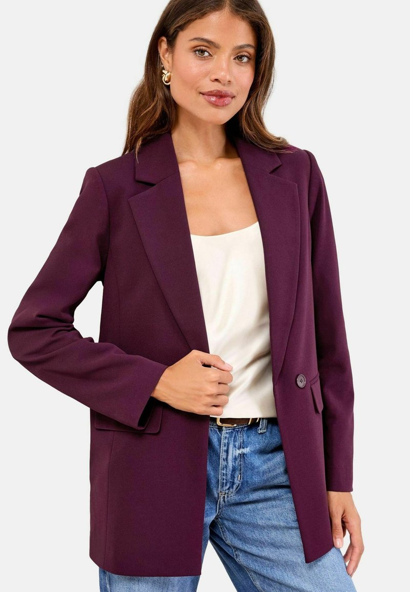 Lilla blazer med et struktureret design, enkelt knaplukning og to forlommer, parret med en cremefarvet top og blå jeans. Silkeagtig tekstur.