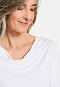 Witte top met gedrapeerde halslijn, zachte stof, gladde textuur, comfortabele pasvorm, waarbij de zijkant van de halslijn en het sleutelbeengebied zichtbaar zijn.