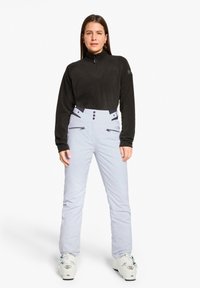 Schwarzer Fleece-Pullover mit Reißverschlusskragen, kombiniert mit hellblauen Schneehosen mit mehreren Reißverschluss-Taschen und Knopf-Details. Schneestiefel inklusive.