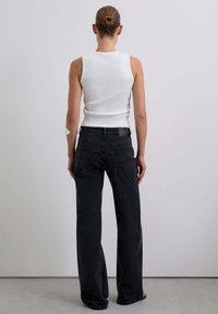 Donna con i capelli raccolti in un chignon, vestita con un top senza maniche bianco e jeans neri larghi, in piedi davanti a una parete bianca uniforme.