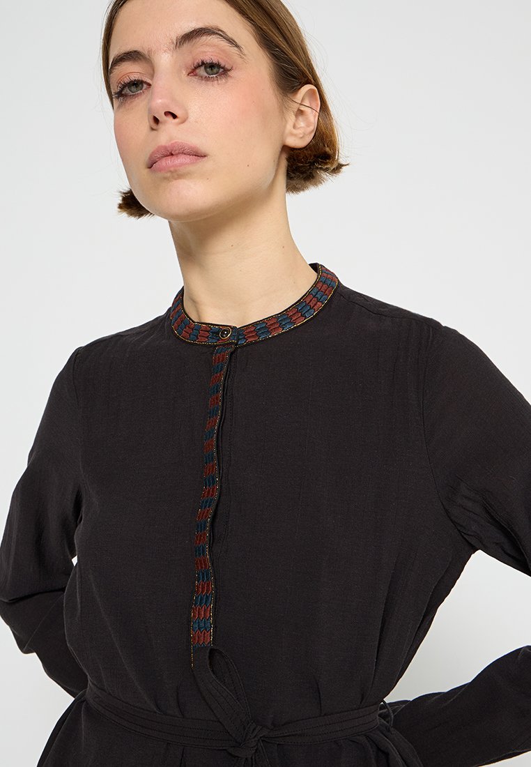 Schwarzes Kleid mit strukturierter Oberfläche, ausgestattet mit einem bunten gemusterten Kragen, einer durchgeknöpften Leiste und einem gebundenen Taillenzug für eine figurbetonte Silhouette.