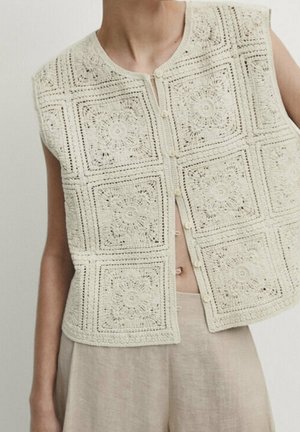 Veste sans manches crochetée avec des motifs floraux carrés, portée sur un pantalon beige clair, avec de petites fermetures à boutons sur le devant.