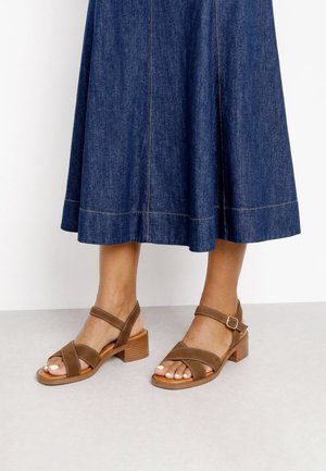 Sandalias de tacón de cuero marrón con tiras en el tobillo, combinadas con una falda midi de denim azul oscuro, mostradas sobre un suelo gris claro.