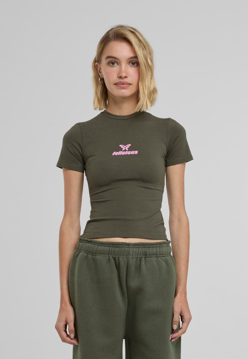 Tricou ajustat de culoare verde măsliniu, cu mâneci scurte, având un design cu fluture roz și text în centrul său. Material moale din bumbac.