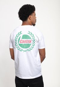 Bílá bavlněná tričko s krátkým rukávem, které na zádech obsahuje zeleno-červený grafický design loga "Castrol" obklopeného vavřínovými listy.