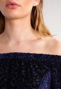 Granatowa bluzka off-the-shoulder z teksturowanym kwiatowym wzorem, zebraną elastyczną linią dekoltu i gładkim materiałem, podkreślającym delikatny połysk.