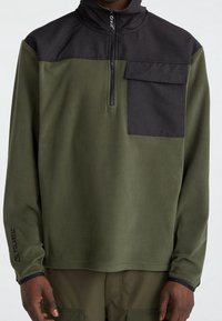 Olijfgroene fleece trui met een zwart bovenpaneel, een halve ritssluiting en een borstzak. Gestructureerde stof met Polartec-logo op de mouw.