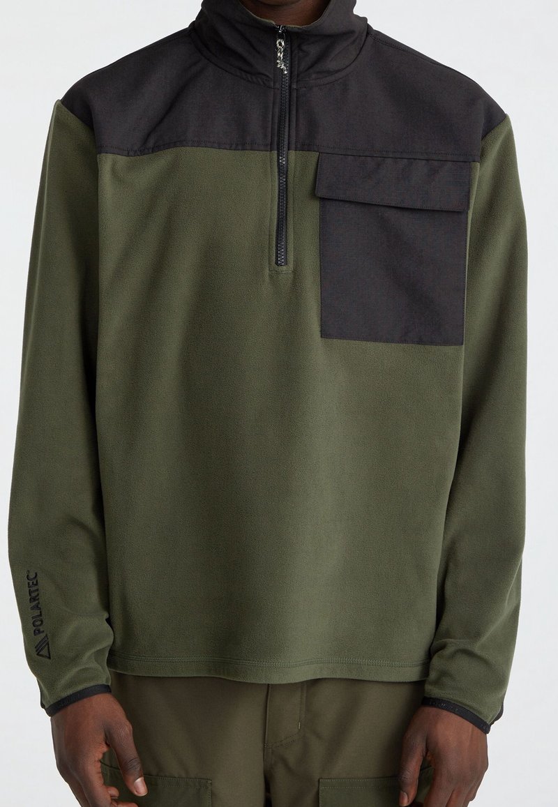 Olijfgroene fleece trui met een zwart bovenpaneel, een halve ritssluiting en een borstzak. Gestructureerde stof met Polartec-logo op de mouw.