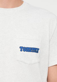 Detailný záber svetlošedej náprsnej kapsy trička s modrým logom "Tommy Jeans" na hornej časti tela osoby.