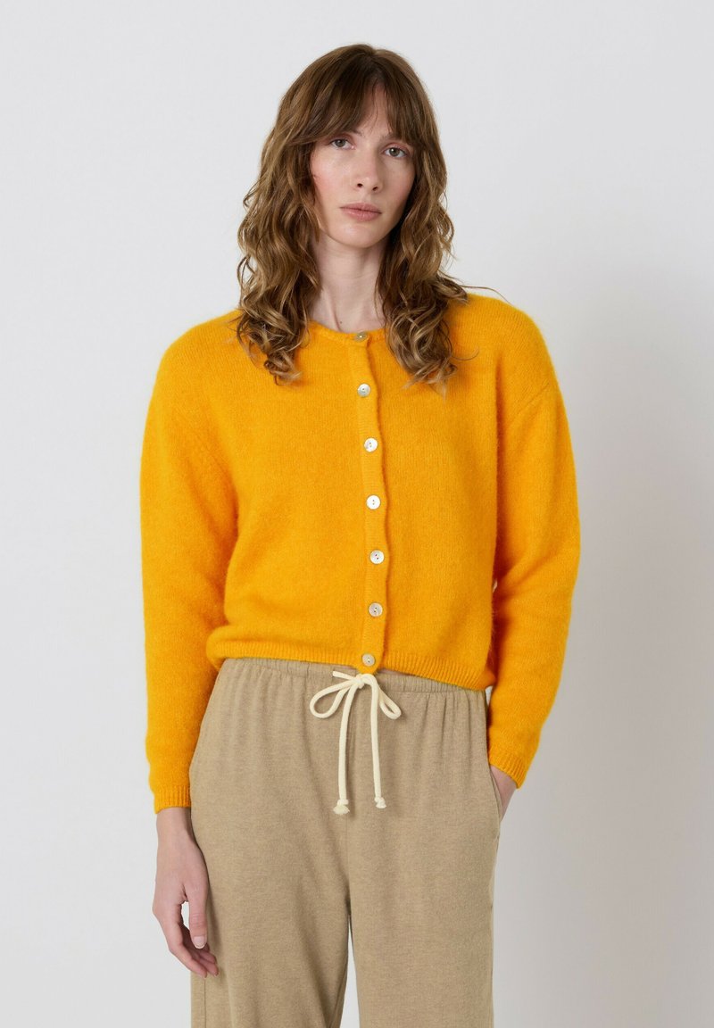 Ryškiai geltonas kardiganas iš minkštos medžiagos, turi apvalų kaklą, priekinę sagų eilę ir ribsčius rankogalius, derinamas su smėlio spalvos kelnėmis su raišteliu.