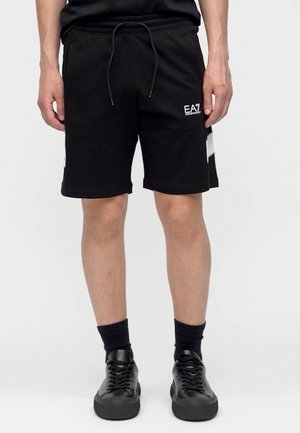 Homme portant un short noir EA7 à cordon, des chaussettes noires et des baskets noires, debout sur un fond blanc uni.