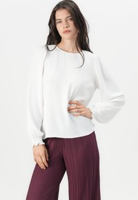 White long-sleeve blouse with a round neckline and gathered cuffs, paired with deep purple pleated trousers, smooth fabric and relaxed fit.