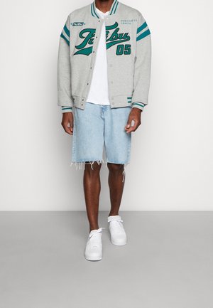 Grå varsityjacka med tealfärgade ränder och bokstäver, kombinerad med ljusblå slitna denimshorts och vita sneakers.