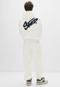 White hoodie featuring a large black script logo on the back, paired with matching white sweatpants and grey sneakers. Smooth fabric texture.