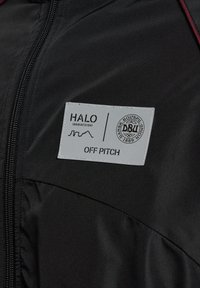 Sort jakke med et tekstureret stof, en lynlås foran og et fremtrædende gråt mærke med teksten "HALO" og "OFF PITCH", samt et logo.