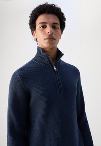 Navyfarbener Zip-Up-Pullover aus geripptem Stoff, mit hohem Kragen und kontrastierender innerer Farbe. Strukturierter, figurbetonter Schnitt.