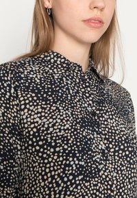 Camicia button-up di un blu navy scuro con un motivo a pois beige e azzurro chiaro. Tessuto morbido con colletto classico e chiusura a bottoni.