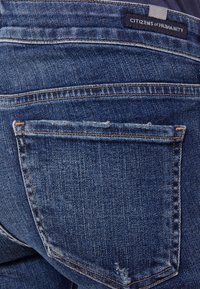 Vaqueros de mezclilla azul oscuro con un diseño clásico de cinco bolsillos, detalles deshilachados en el bolsillo trasero y una etiqueta de la marca visible en la cintura.