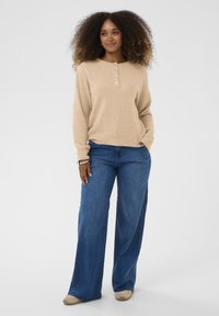 Maglione beige in maglia con scollo henley e quattro bottoni, abbinato a jeans blu a gamba larga. Tessuto morbido e vestibilità rilassata.